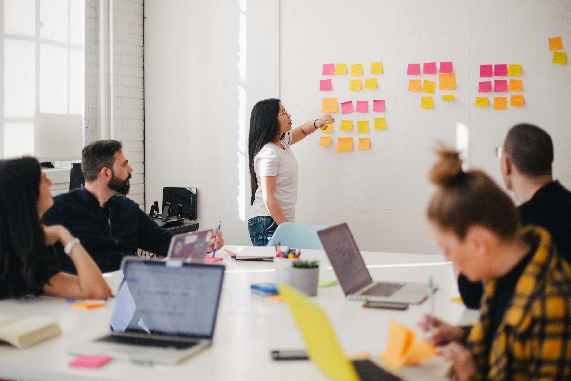 Woman pointing at colourful sticky notes on a wall while colleagues at laptops look on